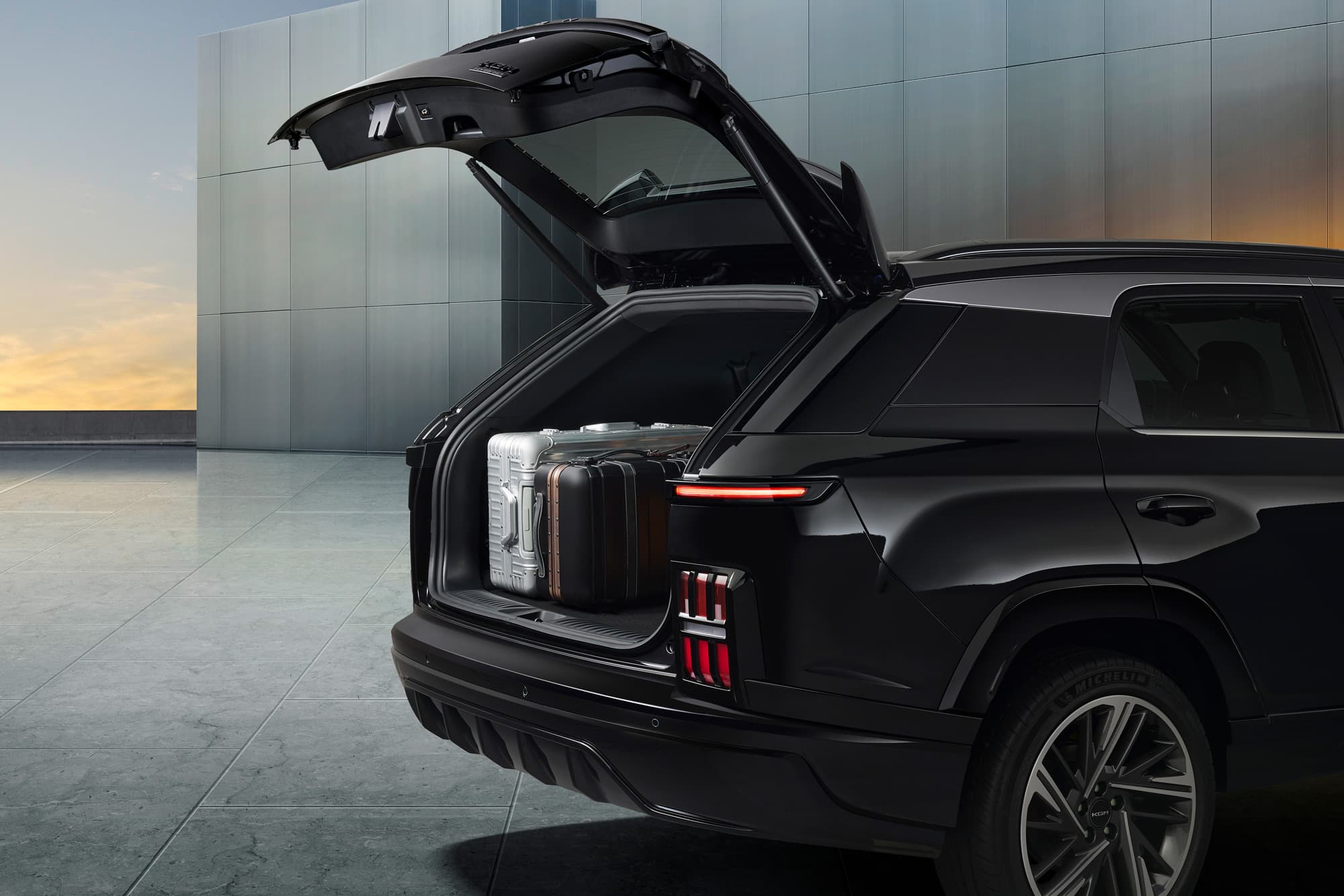 Black KGM Actyon SUV rear with open tailgate showing two suitcases in cargo area, against a modern building at sunset.