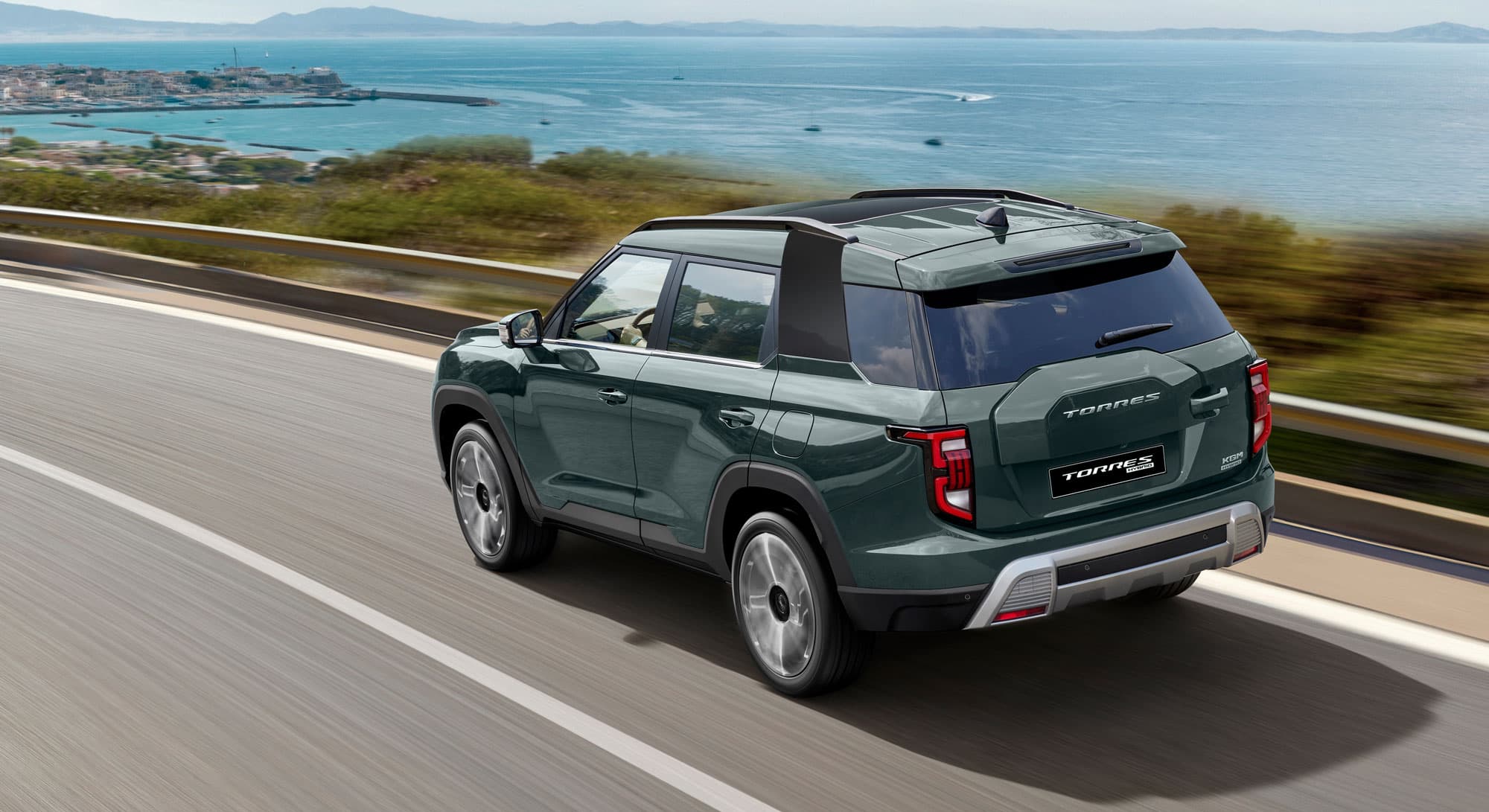 Green  KGM Torres Hybrid driving on a coastal highway with a scenic view of the ocean and distant islands under a clear blue sky.