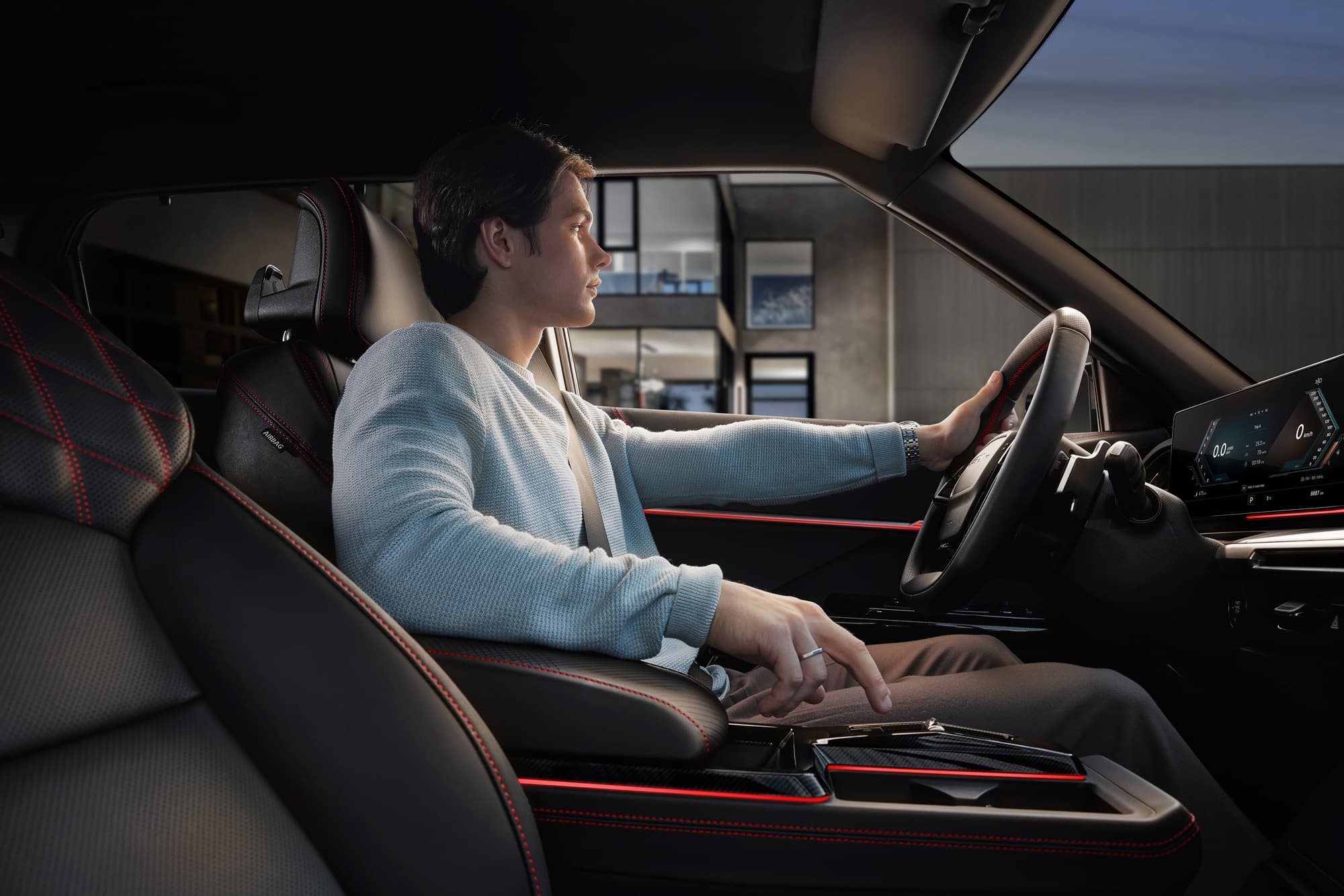 Young man seated in modern car interior, one hand on steering wheel, other on center console, leather seats with red stitching and digital dashboard.
