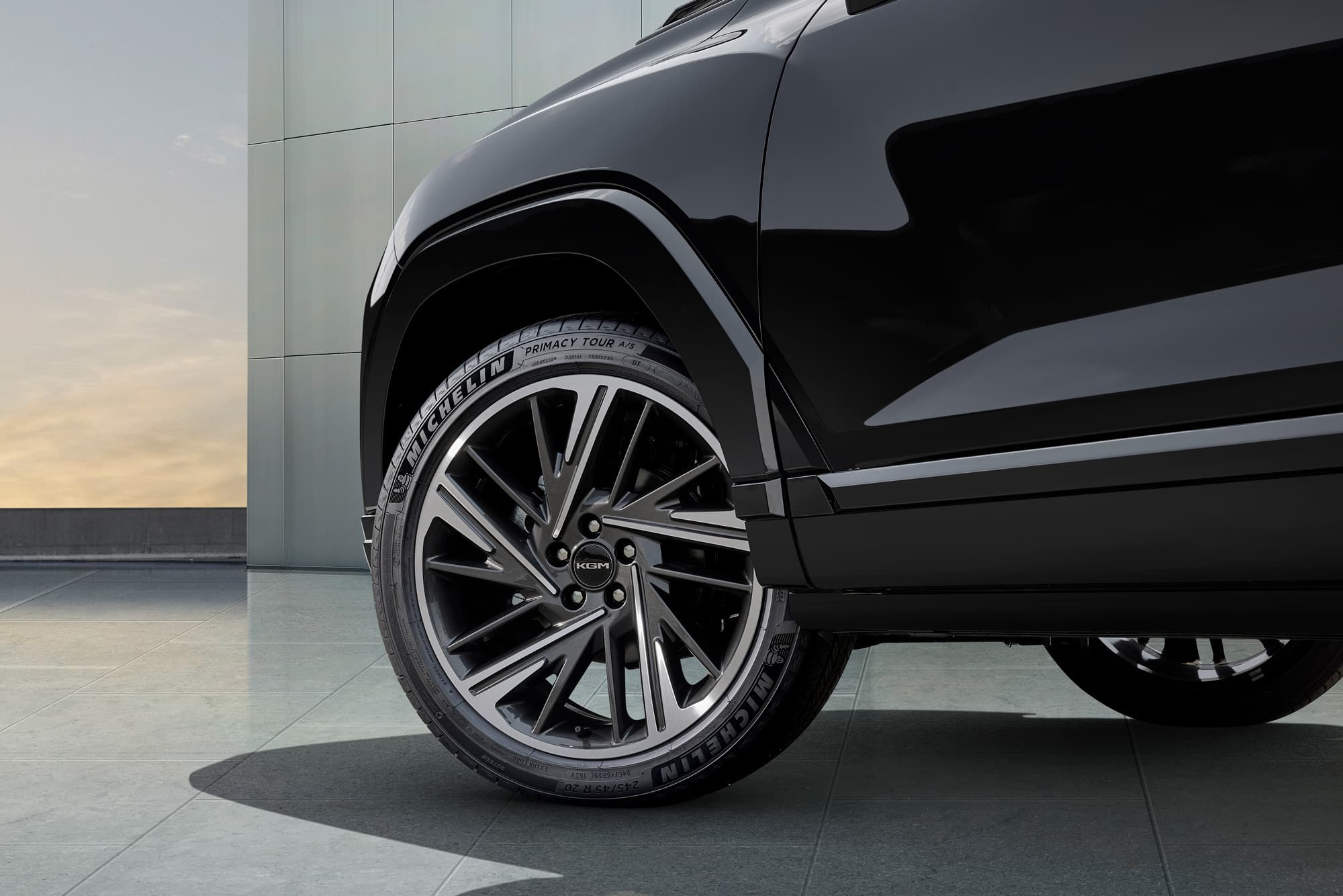 Close-up of black car front wheel with Michelin tire and multi-spoke alloy rim, reflective fender over tiled floor and modern building backdrop.