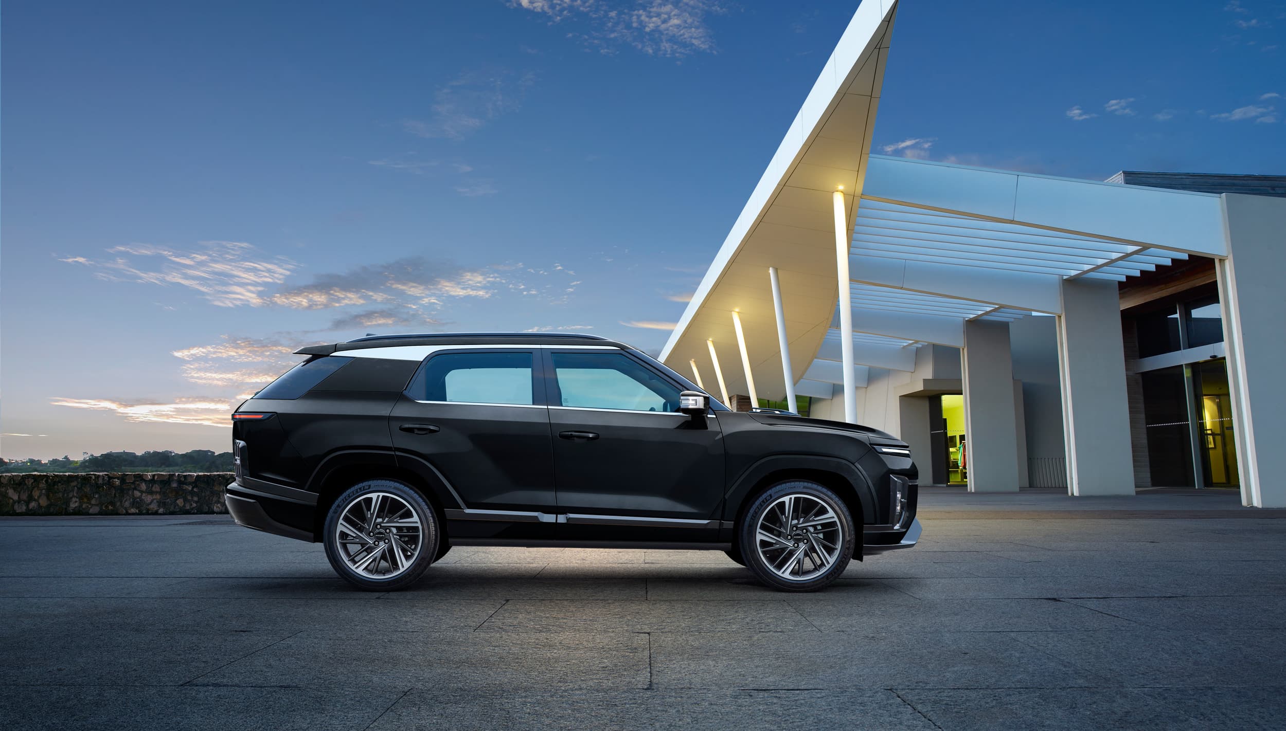 Black KGM Actyon SUV parked by a modern white-canopied building at dusk under a blue sky.