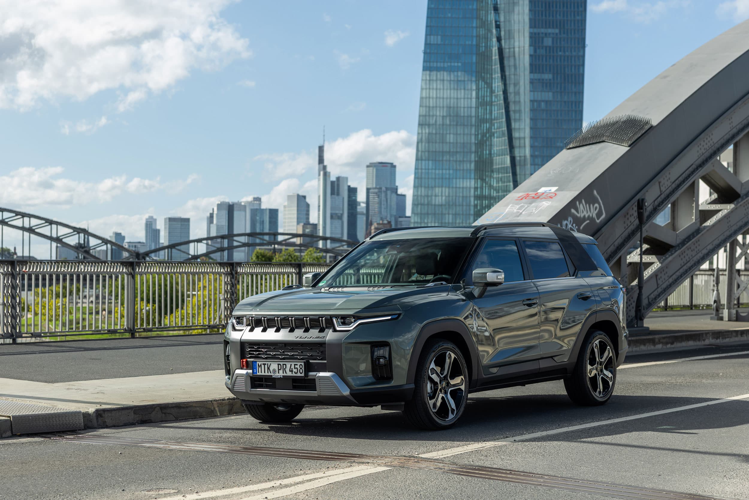 Gray compact KGM Torres Hybrid parked on an urban bridge with metal railings and glass skyscrapers in the background.